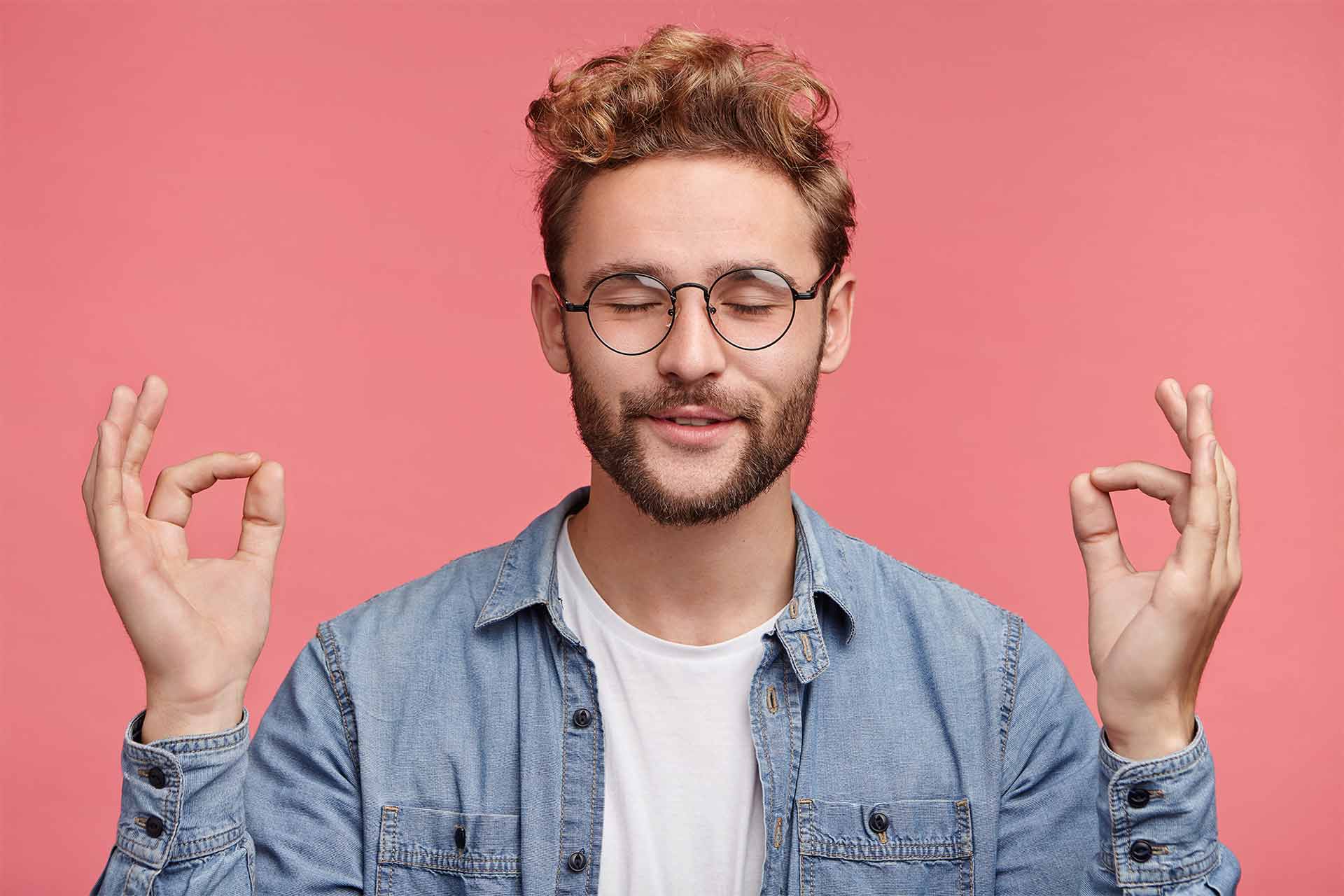 man with white tee shirt and blue collared shirt is relaxed and meditating in front of pink background