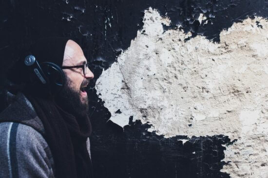 man with glasses wearing black mouth open and a patch of peeling paint next to him making it look like a speech bubble