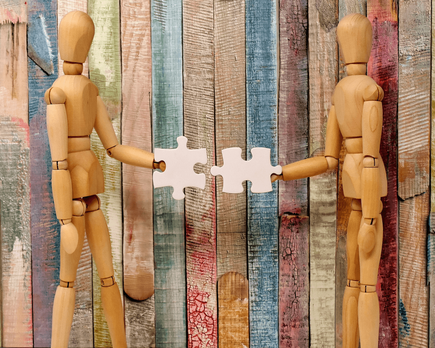 two wooden mannequins for drawing holding two puzzle pieces on a colourful wooden background